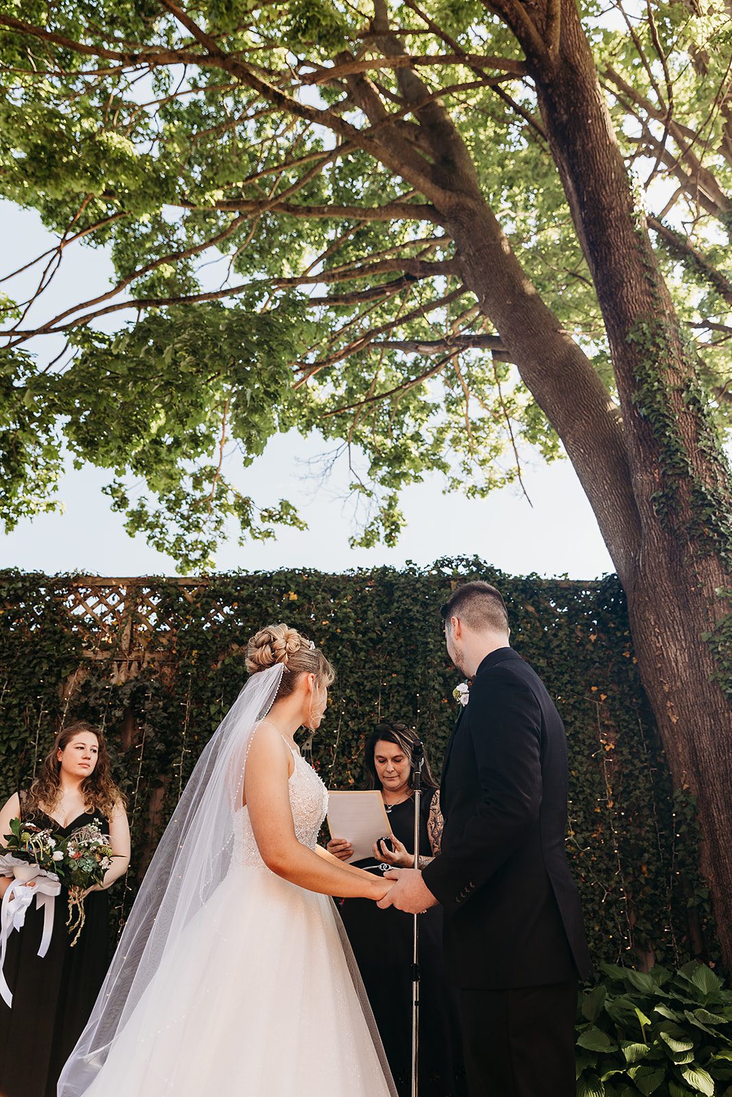 Newlyweds stand in front of their officiant holding hands in a garden under a tree at The Inn At Saratoga Wedding venue
