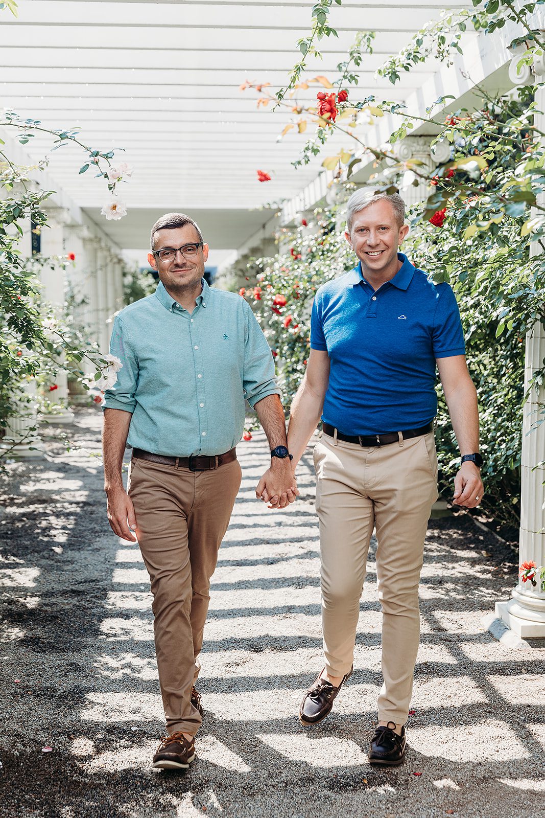 A happy engaged couple holds hands while walking under a rose lined white pergola at the beautiful Yaddo Gardens wedding venue