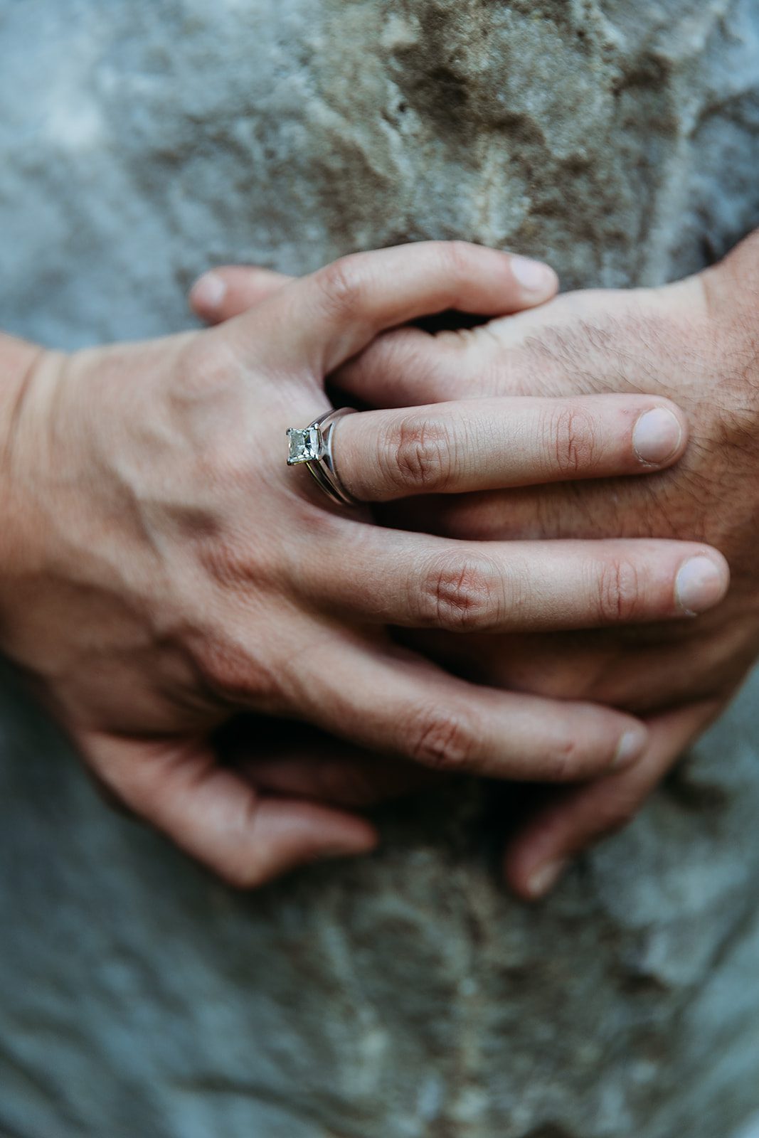 Details of an engagement ring on a hand as it rests on top of another hand on a stone statue