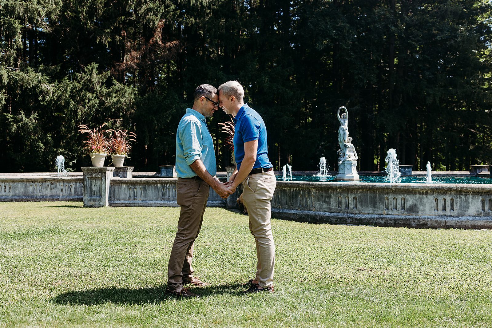 A happy couple touch foreheads and hold hands in front of an ornate fountain in blue polos and khakis