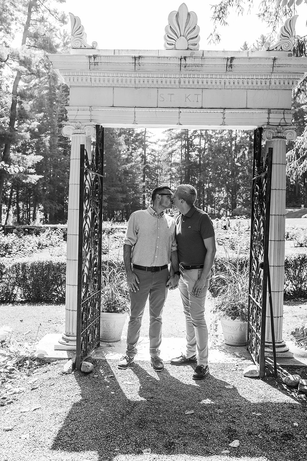 A happy couple holds hands and kisses while standing under an ornate archway and iron gate in a garden