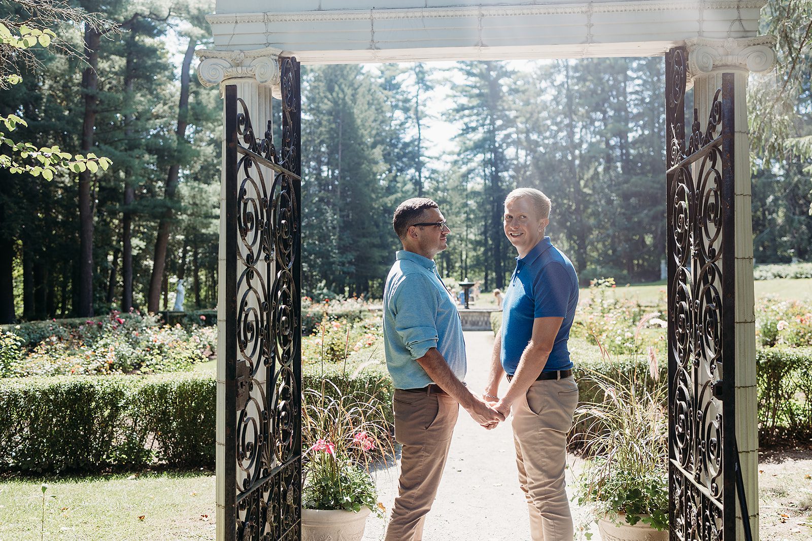 An engaged couple in blue polos and khakis hold hadns and smile under a arch with iron gate at the Yaddo Gardens wedding venue