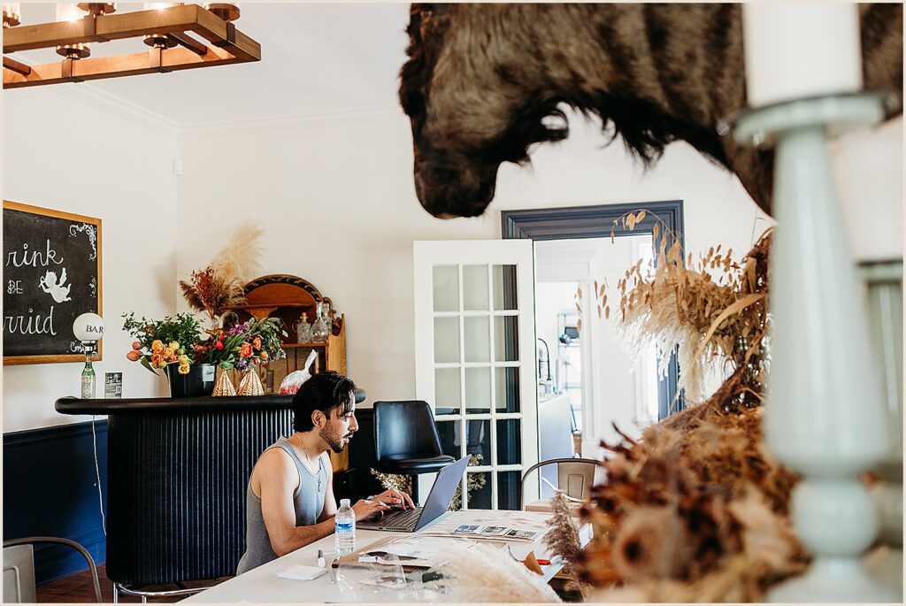 A groom sits at a table and types his vows on a laptop.