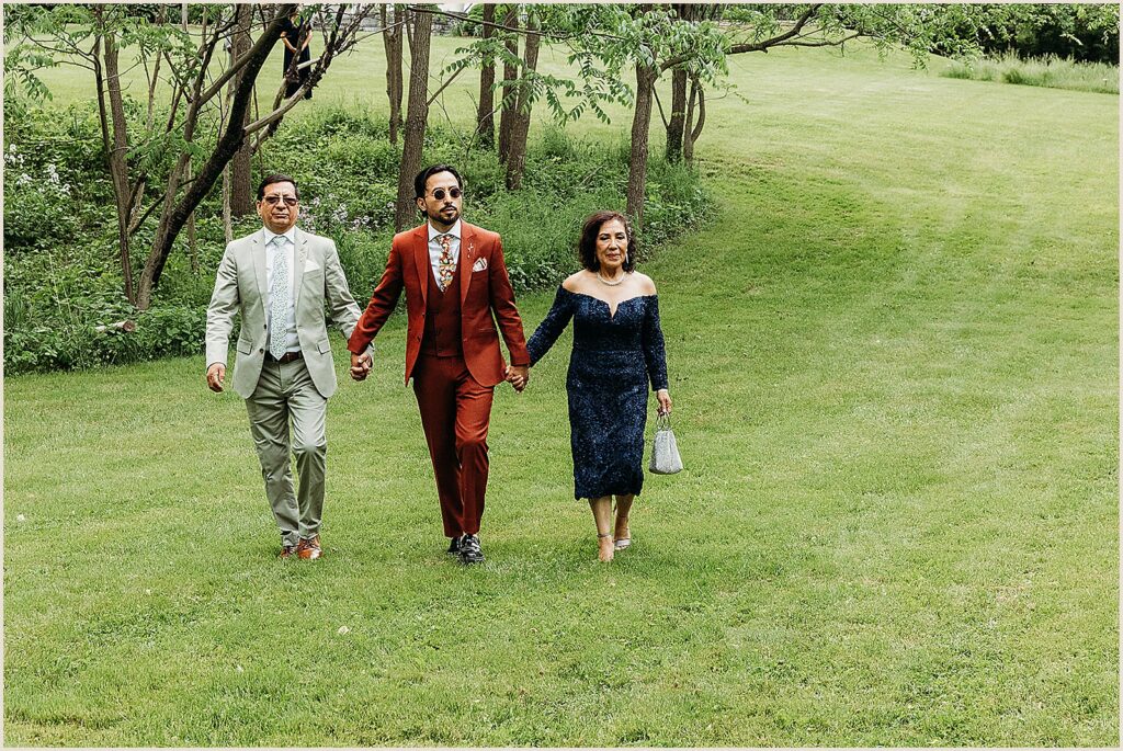 A groom's parents hold his hands as they walk him towards his ceremony.