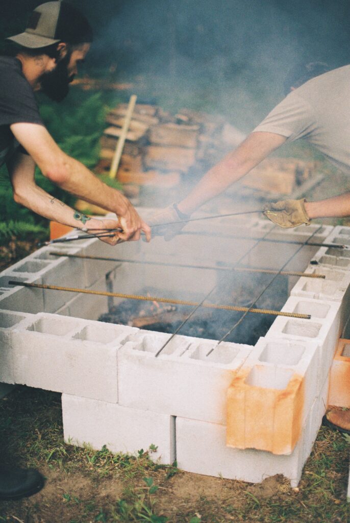 Two men move bars across a roasting pit.