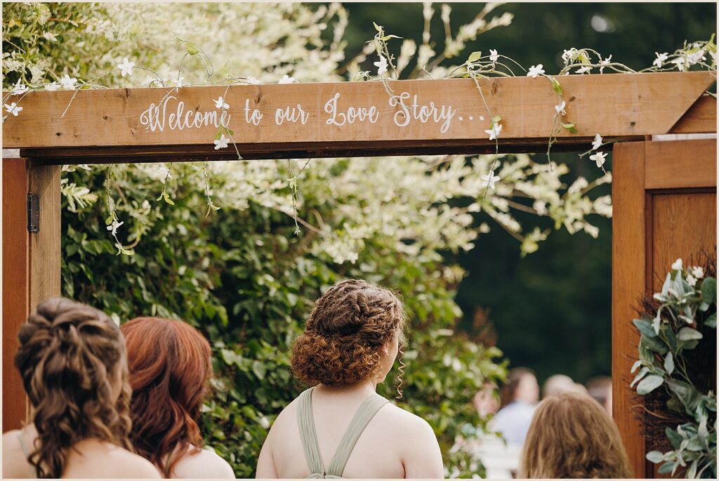Bridesmaids walk under a wooden arch at the start of a wedding ceremony.
