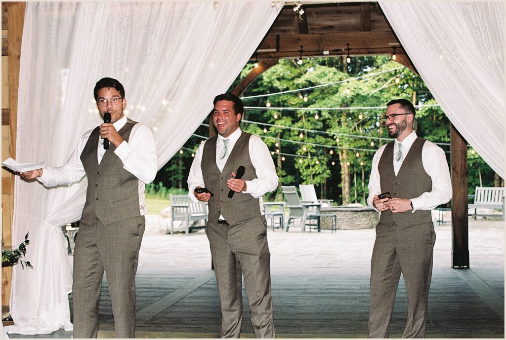 Three groomsmen laugh during a speech in a barn wedding venue.