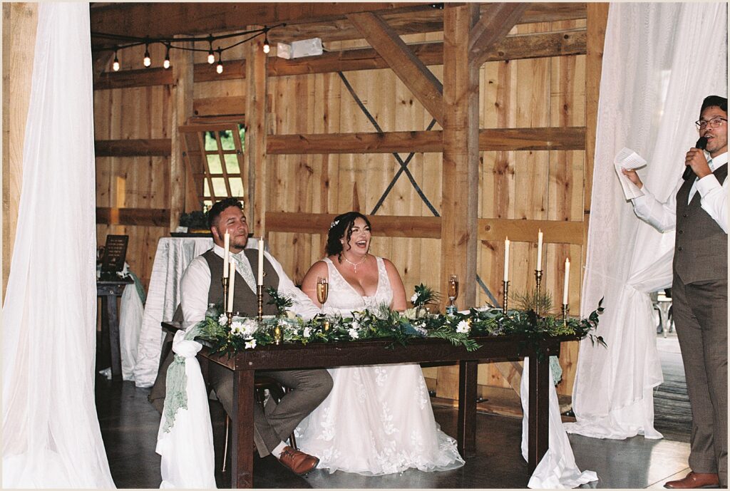 A bride and groom sit at a sweetheart table listening to wedding toasts.
