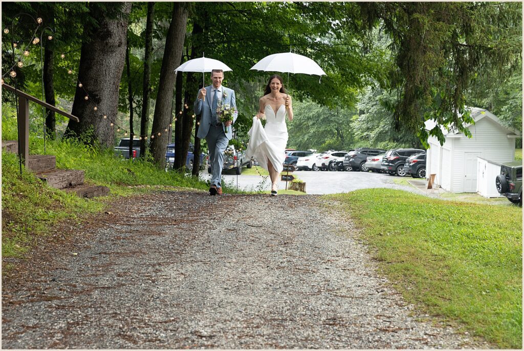 A bride and groom walk down a gravel path carrying umbrellas.