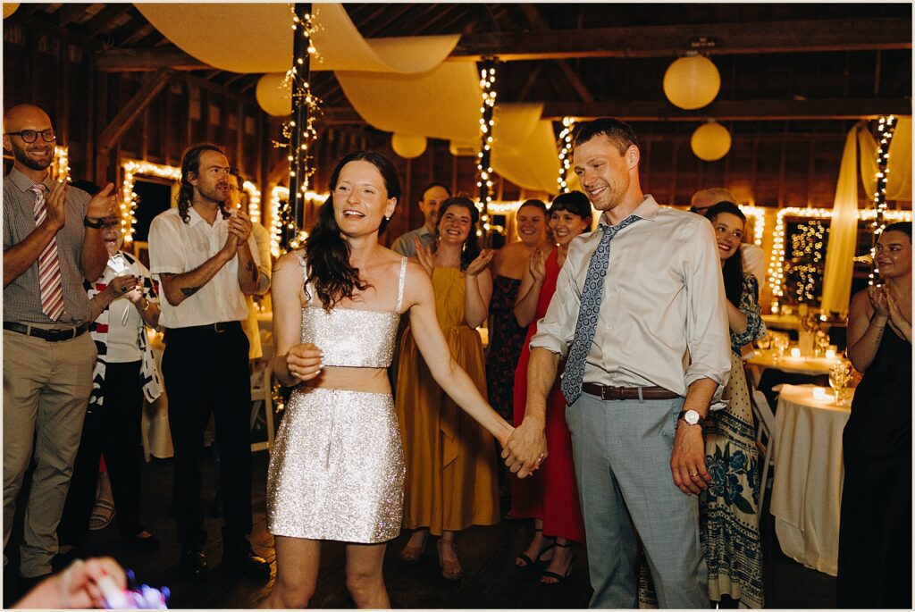 A bride in a sparkly reception dress holds a groom's hand.