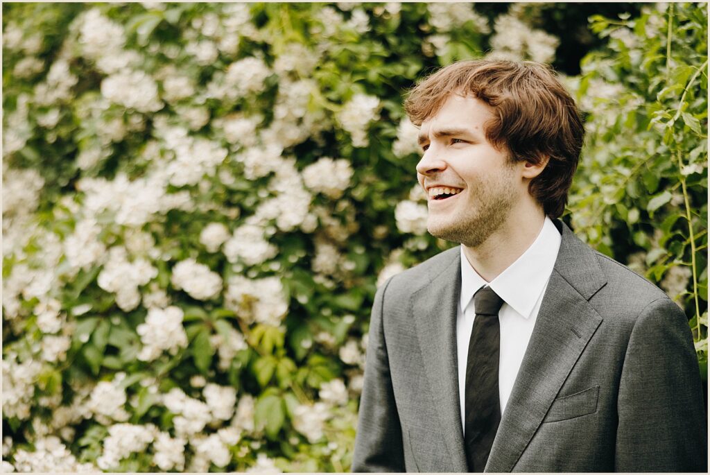A groom poses for an Adirondack wedding photographer in front of a flowering bush.